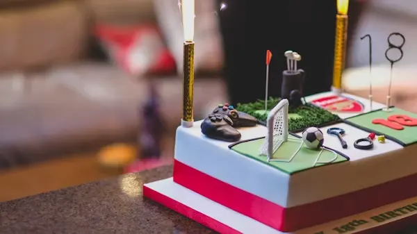 Sports-themed cake with a miniature football pitch, goalpost, golf clubs, game controller and jersey number decorations