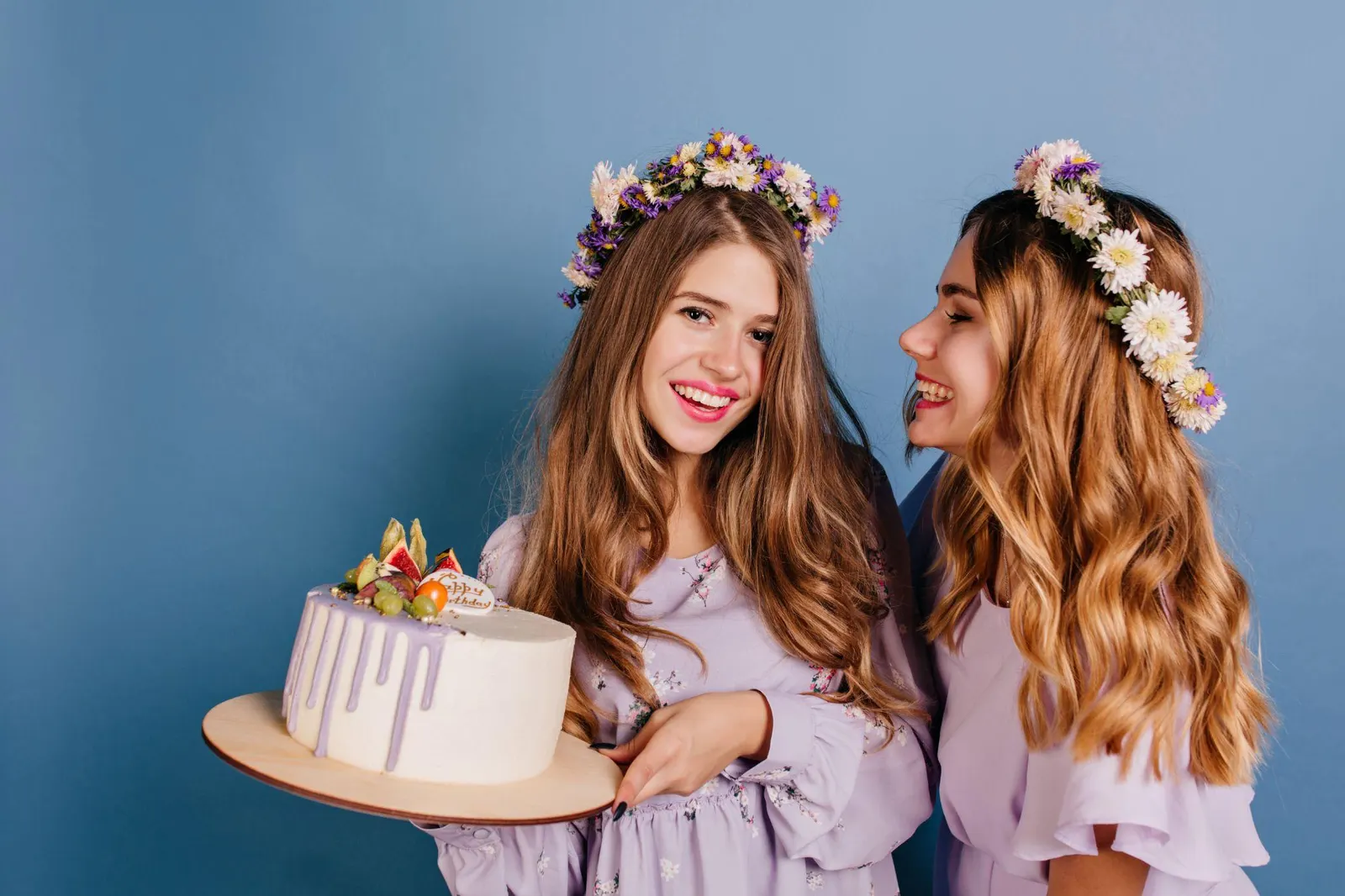 Two friends in flower crowns smiling and holding a white drip cake topped with fresh fruit