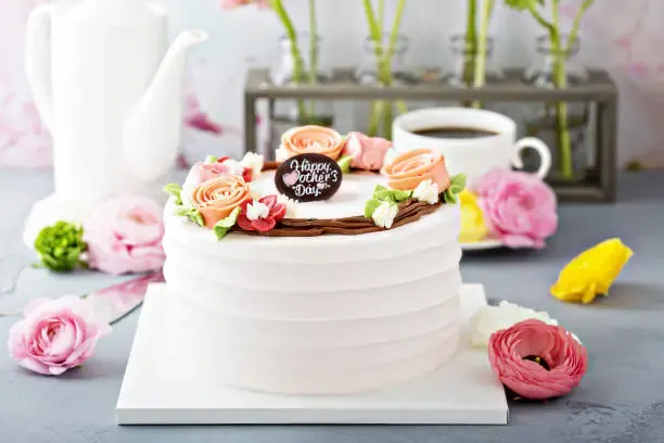 White buttercream cake topped with pink and peach buttercream roses and a 'Happy Mother's Day' plaque, served with coffee and fresh flowers