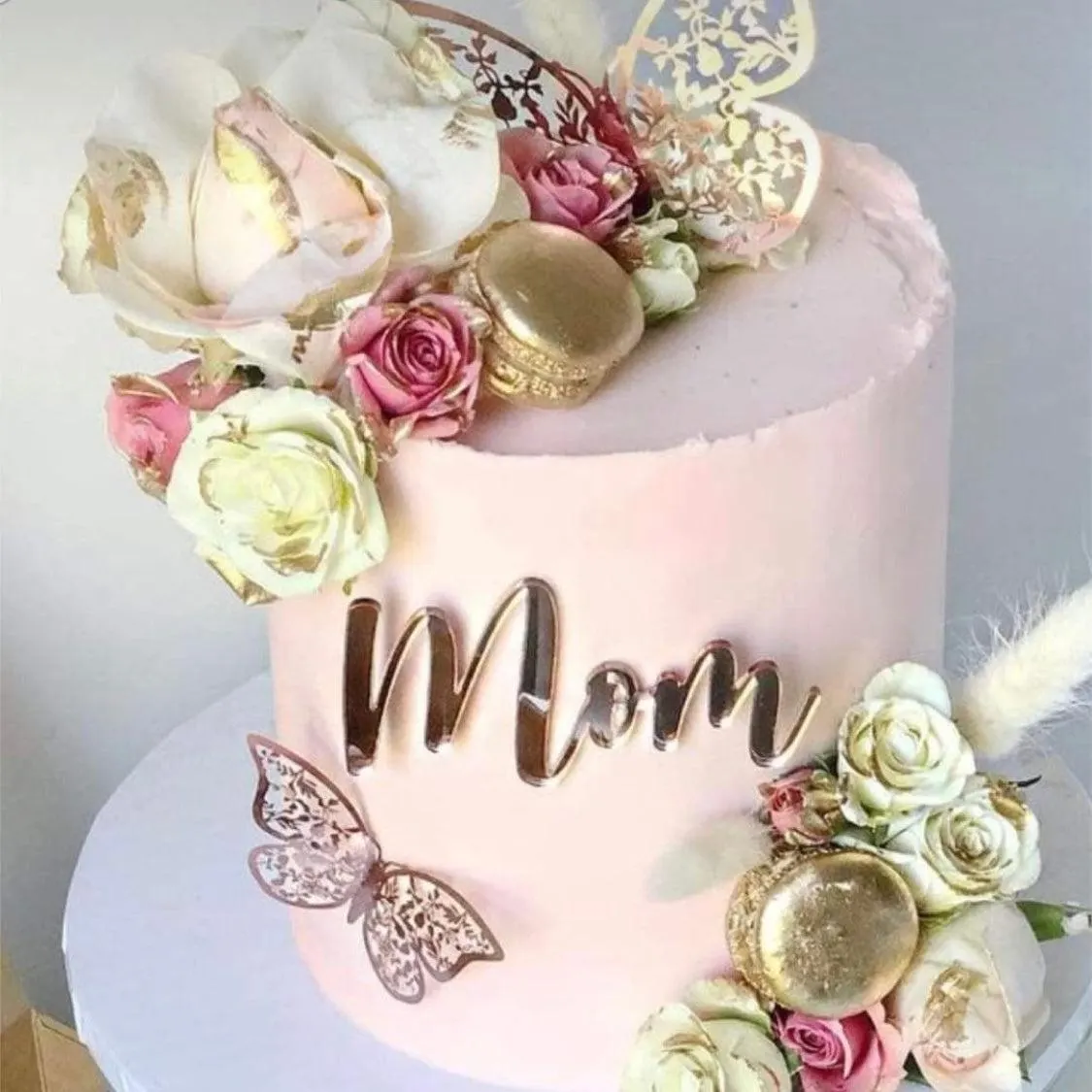 Pink cake decorated with fresh white and pink roses, gold-flecked macarons, a rose-gold 'Mom' topper and paper butterflies