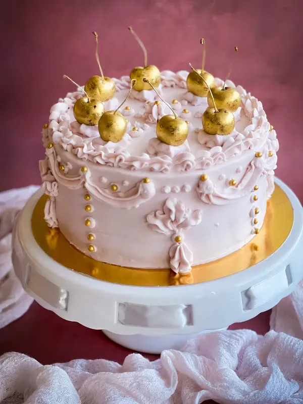 Blush pink vintage buttercream cake with piped scallops, gold beading and gold-leafed cherries on top