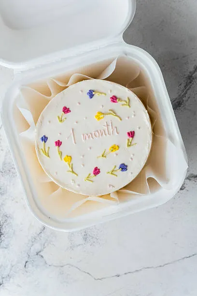 White bento cake hand-piped with flowers and a 1 month message inside an open takeaway box