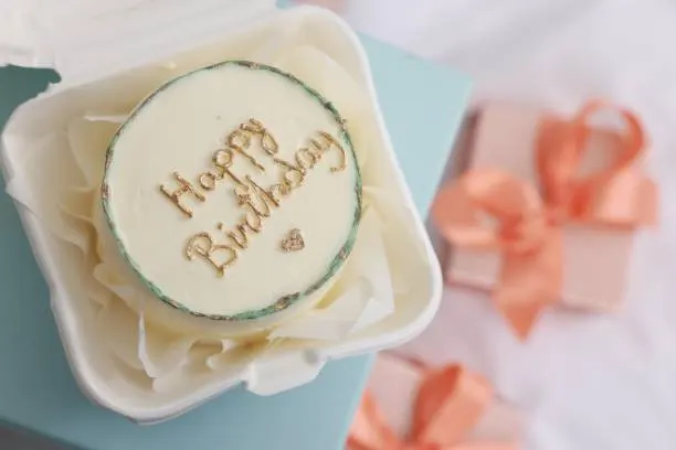 Cream bento cake piped with Happy Birthday in gold script, resting on a mint box beside gift boxes with peach ribbon