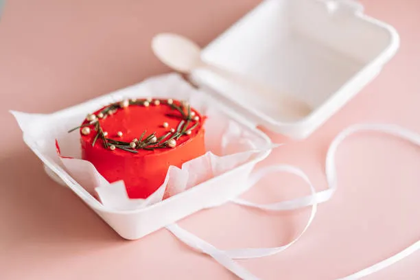 Red bento cake with a gold wreath in a pink takeaway box with a wooden spoon and ribbon