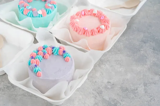 Three pastel mini bento cakes in teal, pink and lavender inside takeaway boxes with wooden spoons